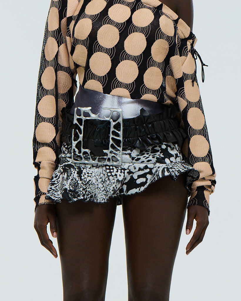 Model wearing a ruffled black leather belt with a very large silver aluminium frame shaped belt buckle on a plain background