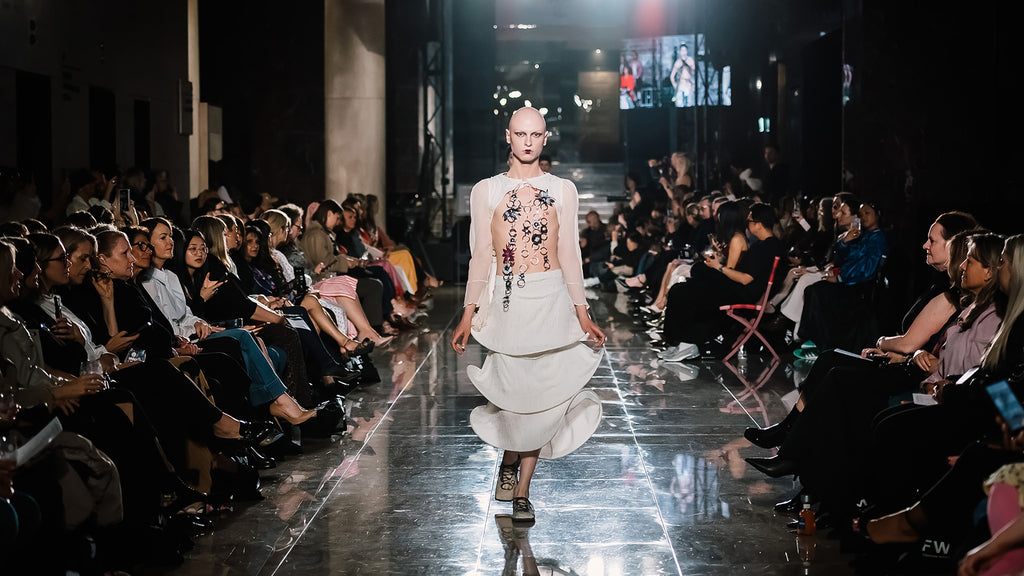 bald model in sculptural white outfit with floral details on chest, walking on a runway at a fashion show with an audience on either side.