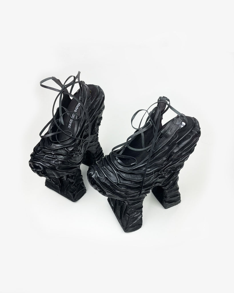 A black, uniquely designed chopine platform shoe with a rippling skin-like leather surface, featuring an open heel and lacing over the front.