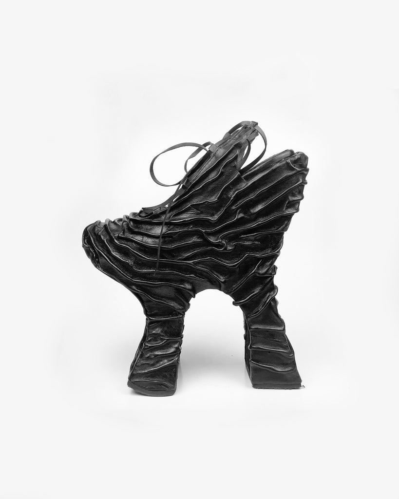 A black, uniquely designed chopine platform shoe with a rippling skin-like leather surface, featuring an open heel and lacing over the front.