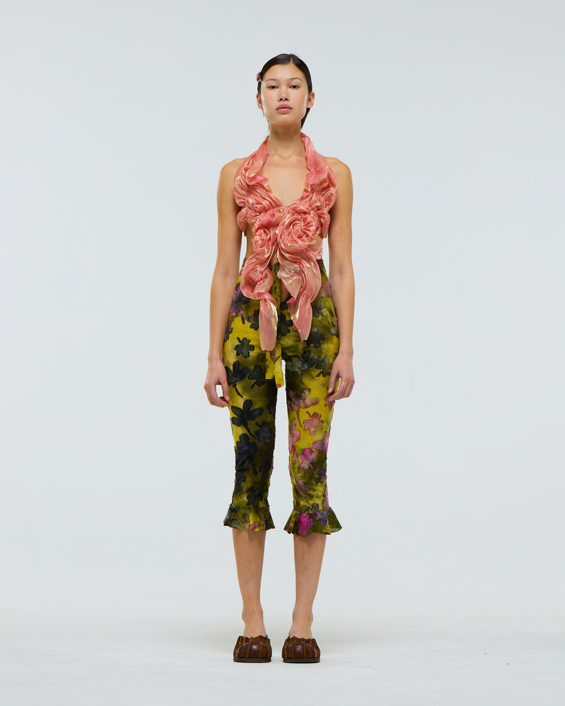 Model wearing a peach coloured ruffled organza top and floral green tie dye  capri pants with frills at the calves on a white background