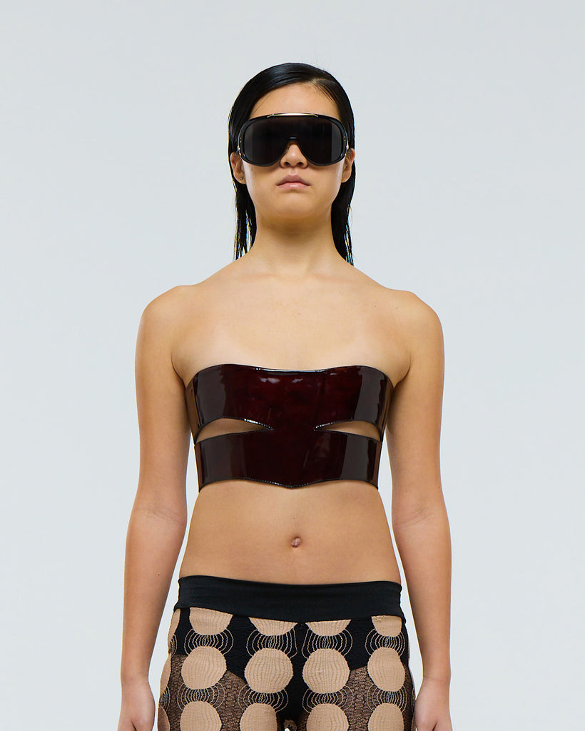Model wearing a dark red burgundy patent leather strapless lace up top and patterned pants on a plain background