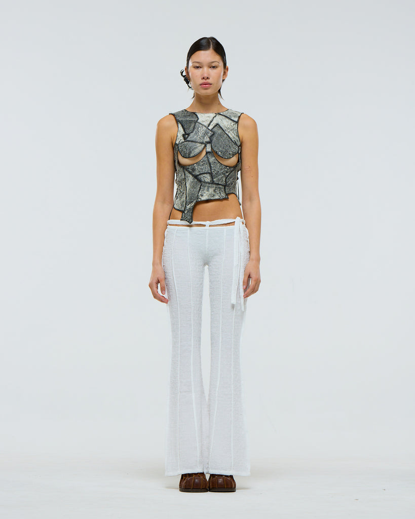 Model wearing a grey and black patterned top with exposed chest areas and white pants with suspended drawstring on a plain background