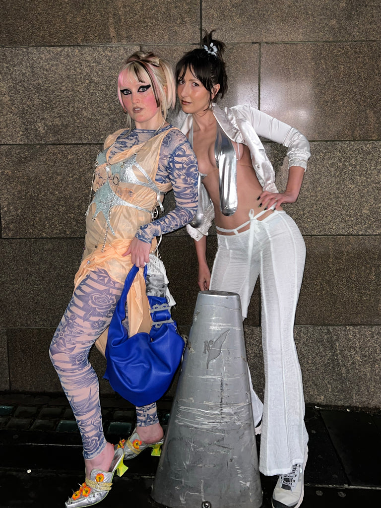 Two women in avant-garde outfits posing against a textured wall.