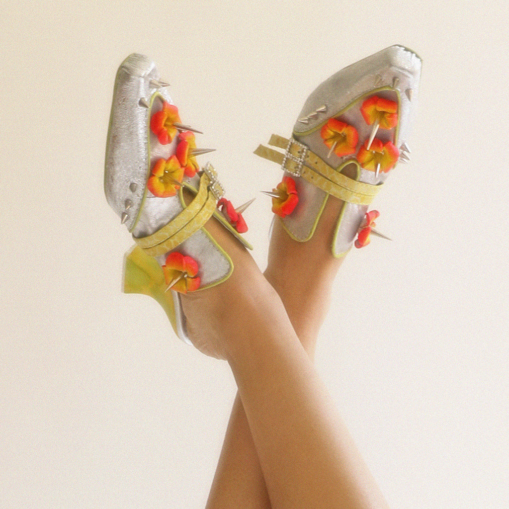 Silver high-heeled shoes with red and orange flower details, silver spike studs and yellow straps with rhinestone buckles on a beige background