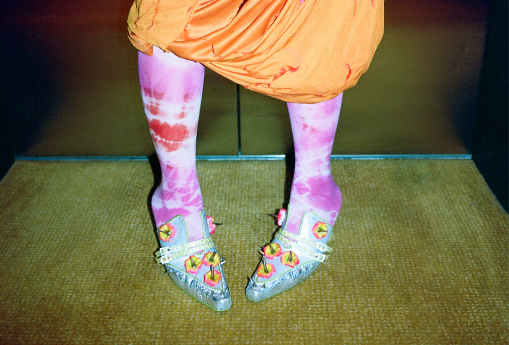 model in orange dress and pink tights, wearing silver shoes on green carpet and green backgroud