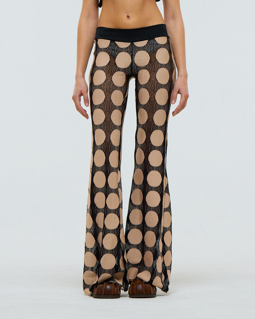 Sheer flared black pants with a beige polka dot pattern worn by a model on a white background
