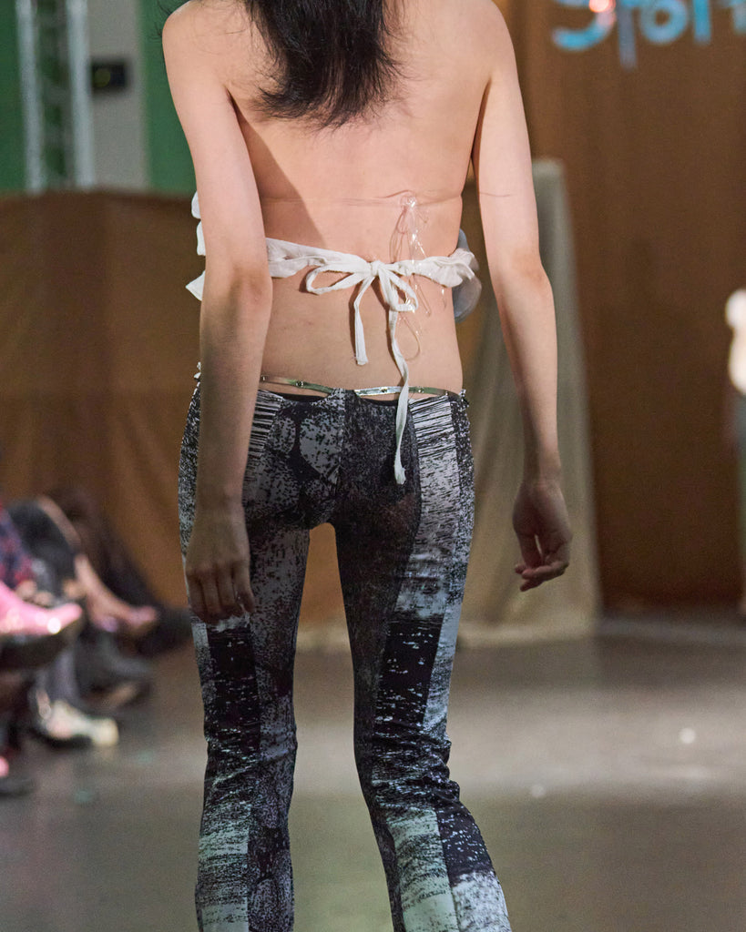 model wearing a pair of black and white very low waist stretch flare pants with a geometric abstract pattern, and super thin silver buckle belt viewed from the back