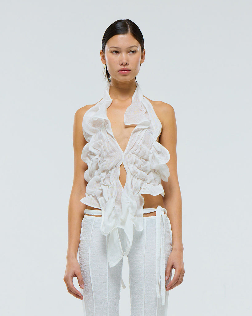 Model wearing a white ruffled linen top and white pants against a light gray background