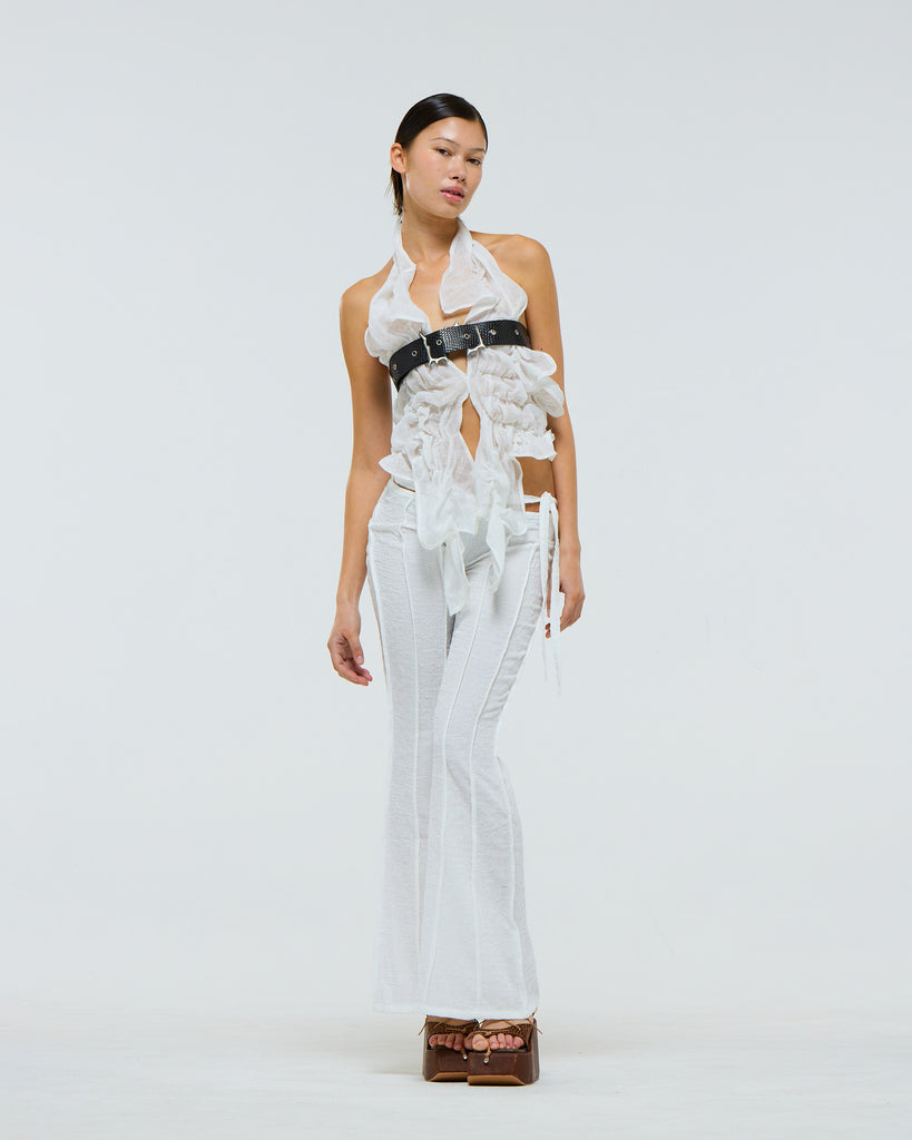 Model wearing a white ruffled linen top with black belt over the chest and white pants against a light gray background