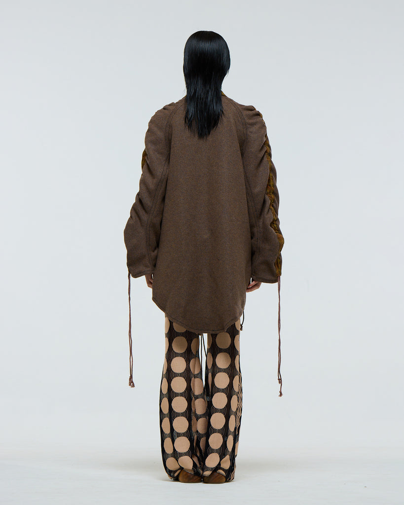 Model wearing a gathered sleeve brown wool jacket with plaid detailing and drawstrings, dark red patent leather corset, and patterned pants on a white background viewed from the back