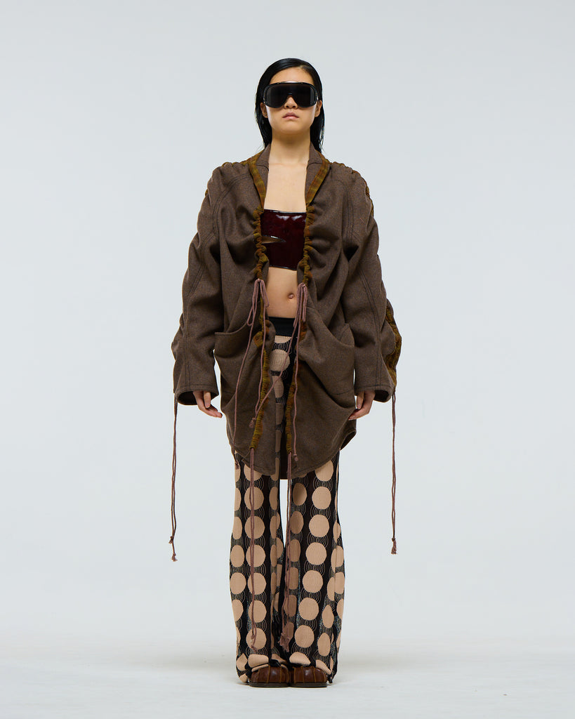 Model wearing a gathered sleeve brown wool jacket with plaid detailing and drawstrings and large pockets, dark red patent leather corset, and patterned pants on a white background