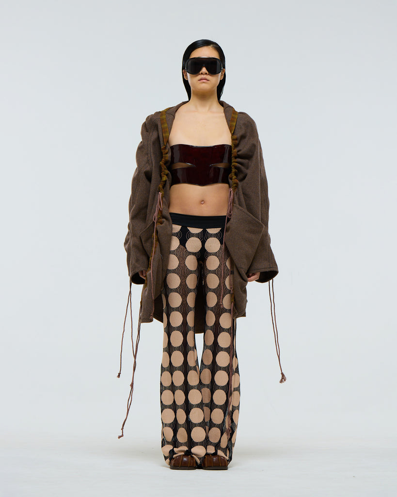 Model wearing a gathered sleeve brown wool jacket with plaid detailing and drawstrings, dark red patent leather corset, and patterned pants on a white background