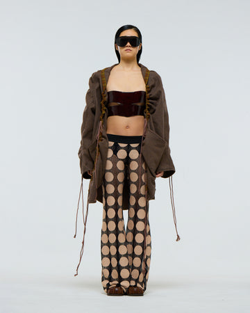 Model wearing a gathered sleeve brown wool jacket with plaid detailing and drawstrings, dark red patent leather corset, and patterned pants on a white background