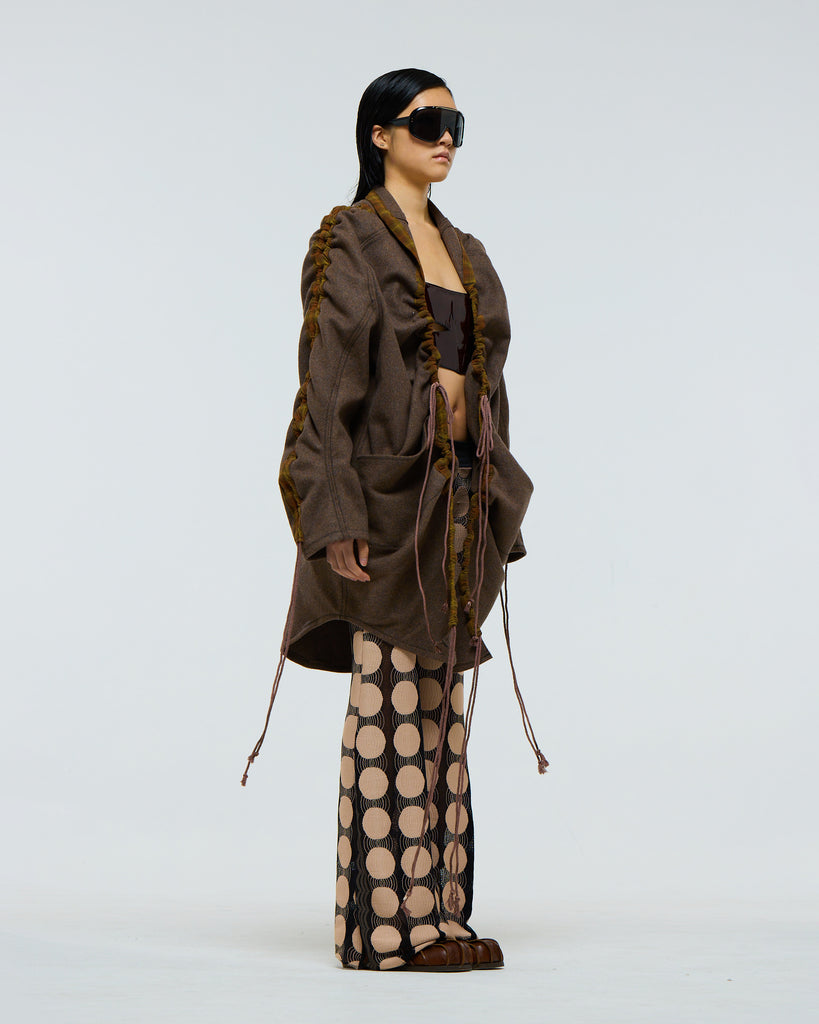Model wearing a gathered sleeve brown wool jacket with plaid detailing and drawstrings, dark red patent leather corset, and patterned pants on a white background viewed from the side