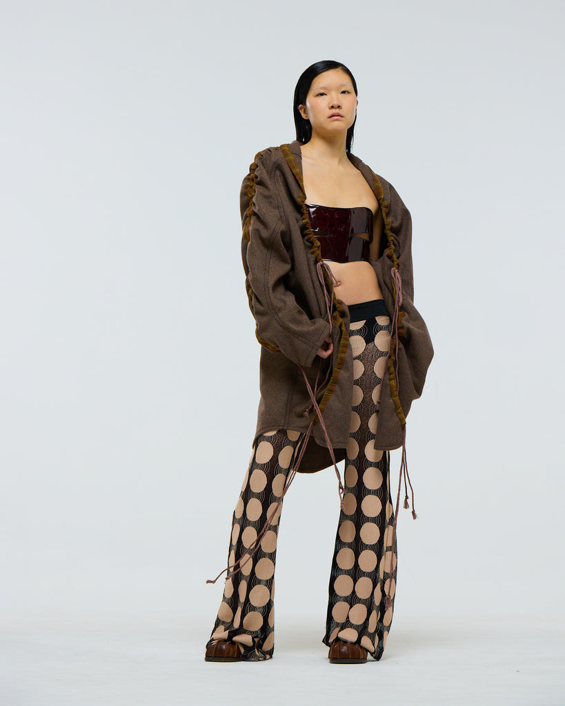 Model wearing a gathered sleeve brown wool jacket with plaid detailing and drawstrings, large pockets and dark red patent leather corset, and patterned pants on a white background