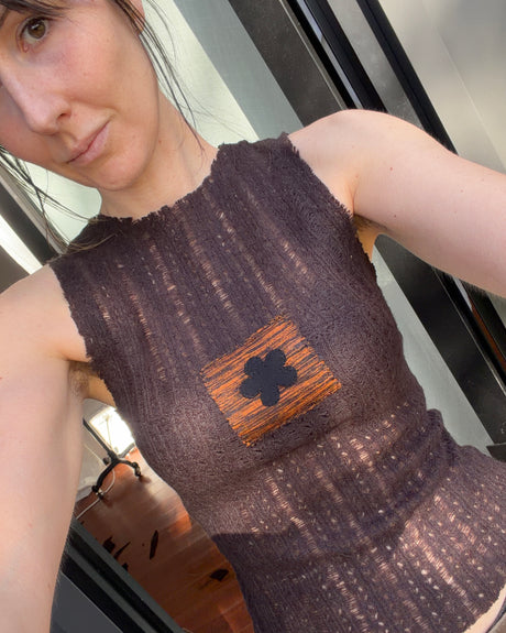 A close-up selfie of a woman wearing a sleeveless, dark brown textured knit top with a square orange-brown patch featuring a black flower on the chest.